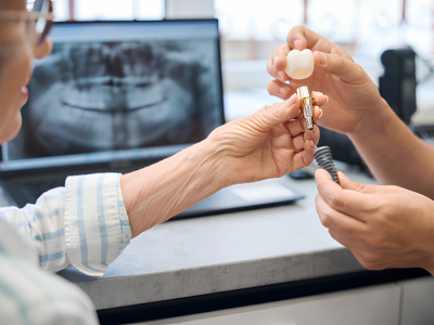 Dentist and patient exchange a dental implant crown and abutment, with a dental X-ray on a computer screen in the background.